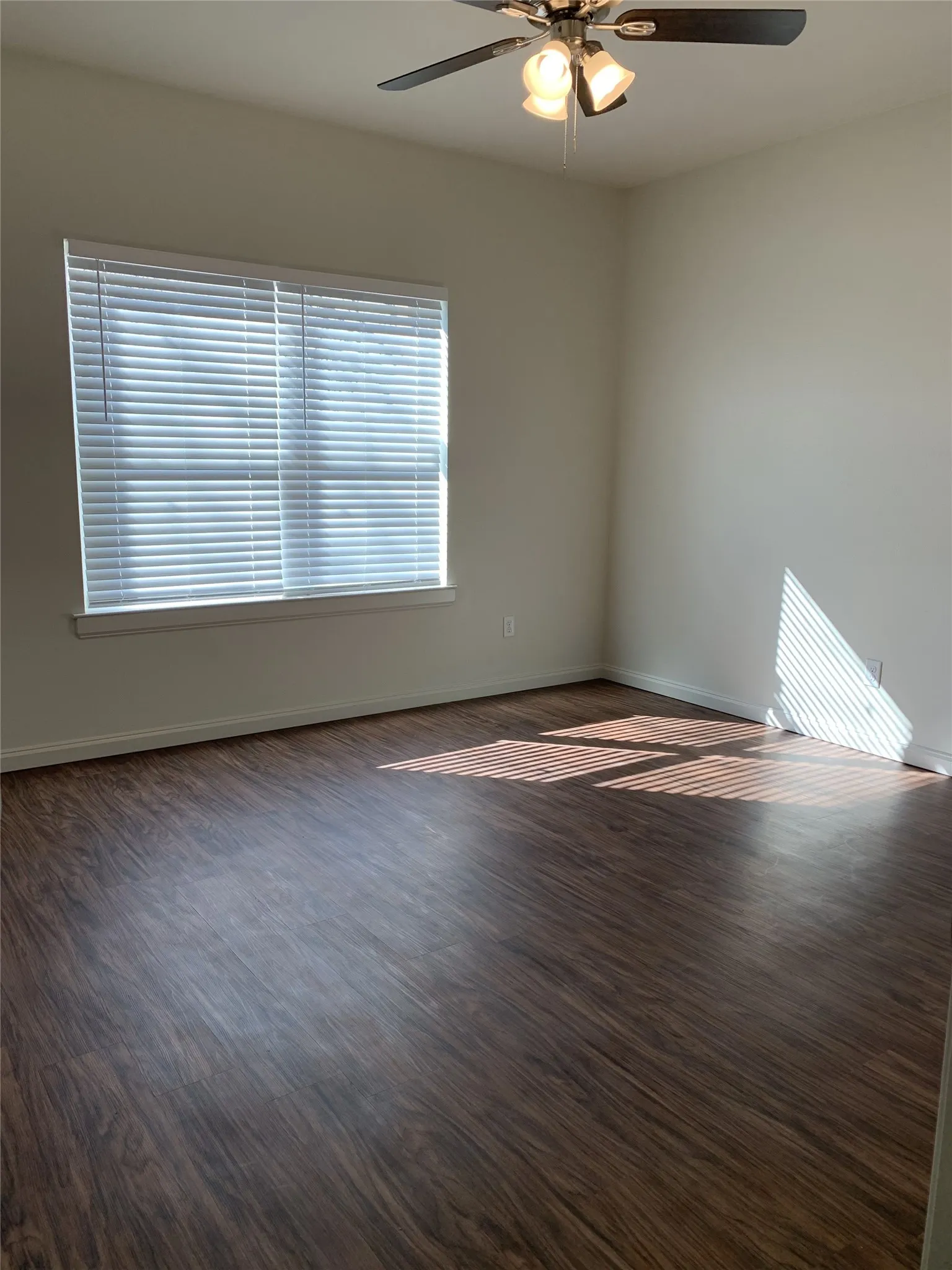 Spacious room featuring wood-finish flooring, a large window with blinds, a ceiling fan with integrated lighting, and neutral wall paint