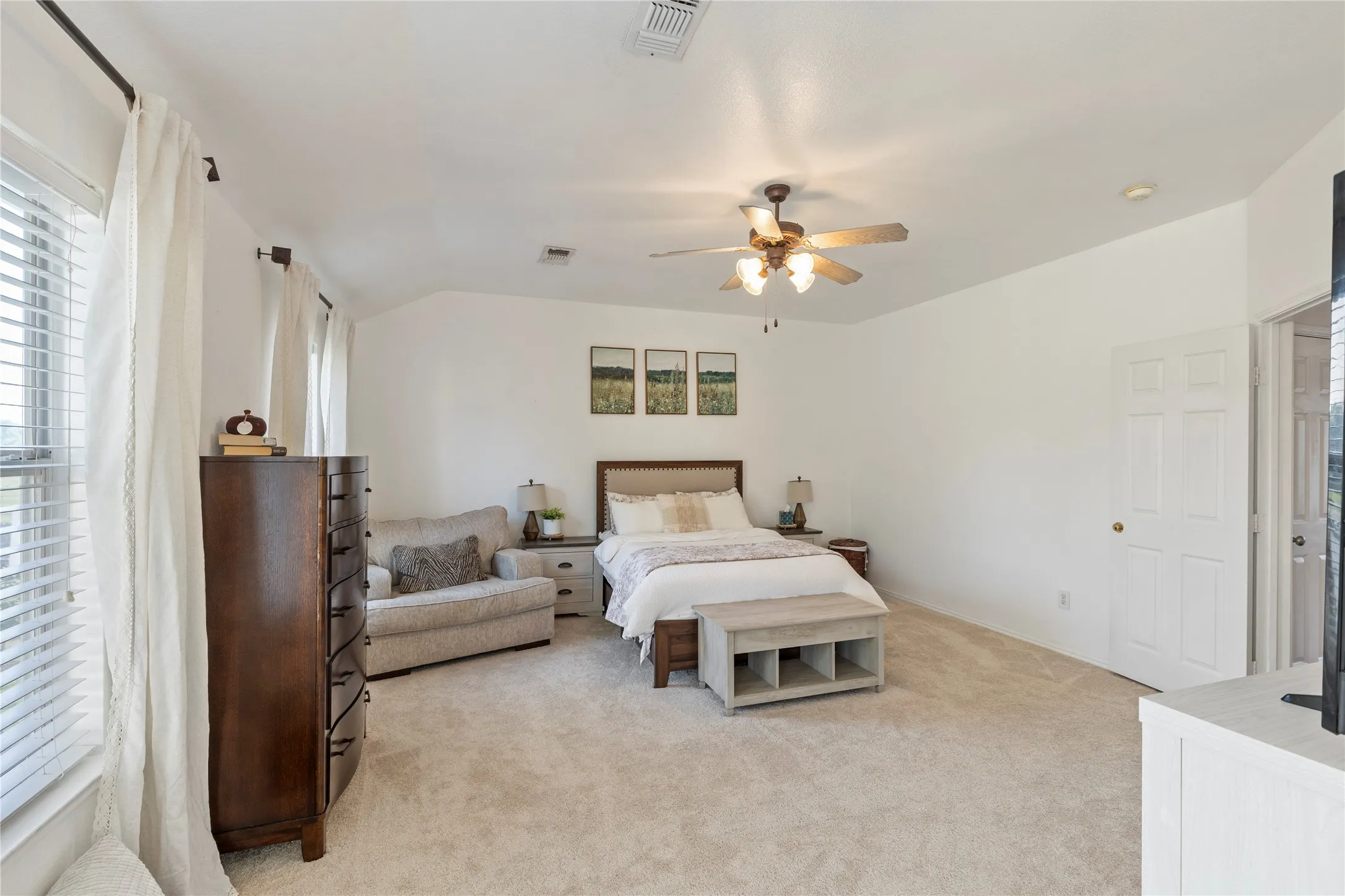 Spacious bedroom featuring plush carpeting, bright white walls, multiple windows with blinds, a ceiling fan with integrated lighting, and a paneled interior door