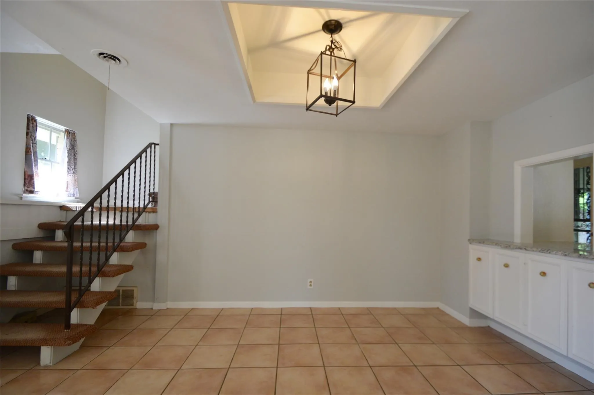 Warm tile flooring throughout, featuring a staircase with black iron railing, a built-in cabinetry unit with a light-colored countertop, a recessed ceiling detail with a suspended lantern fixture, and a pass-through opening