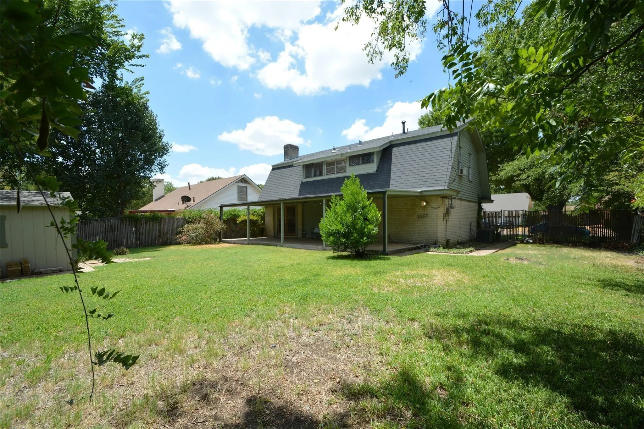 Expansive backyard featuring a covered patio, mature perimeter trees, a storage shed, a wood fence, and a sloped roofline on the main residence