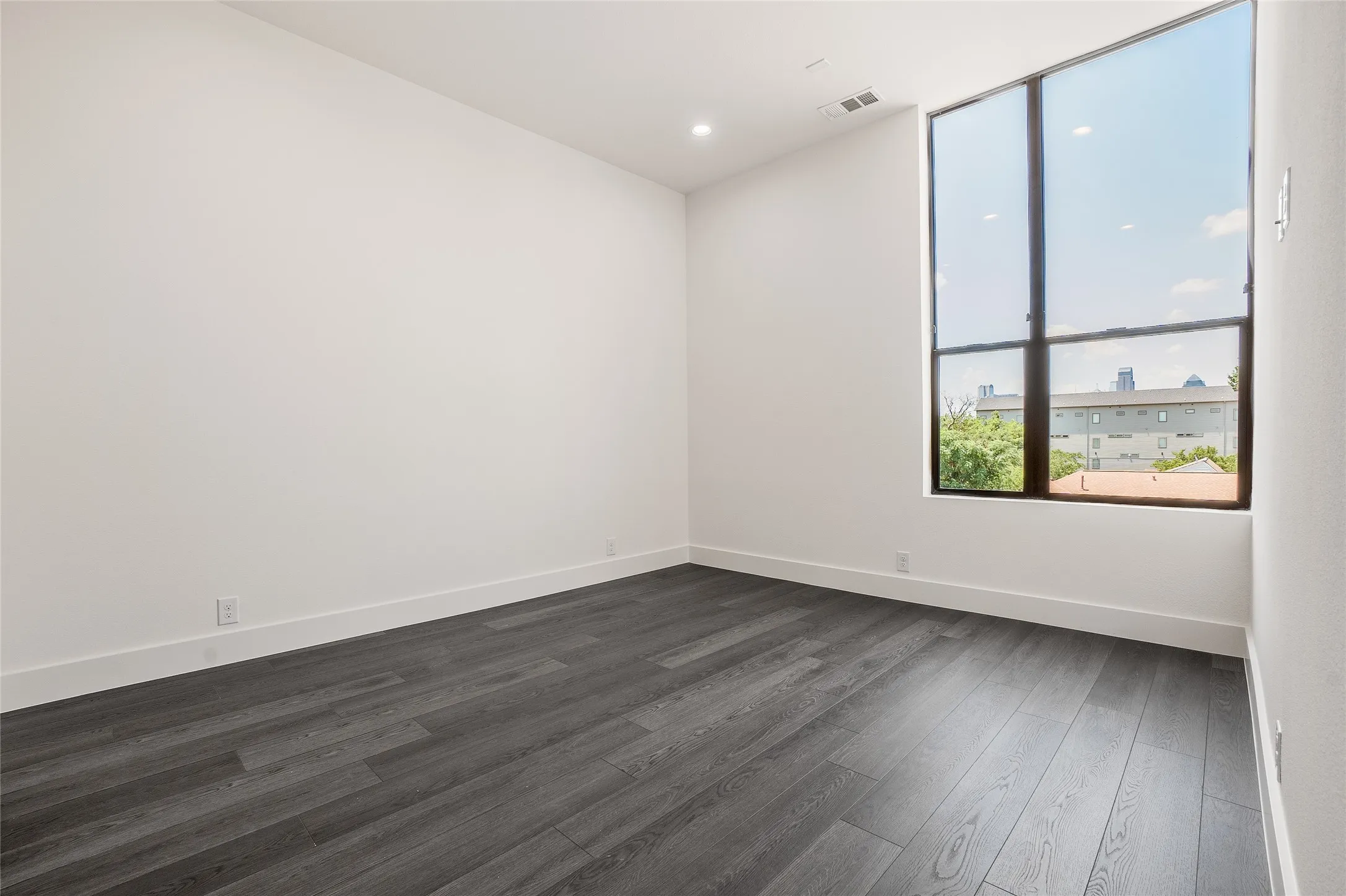 Spacious room featuring a large, multi-paned window, dark wood-finish flooring, white baseboards, and recessed lighting