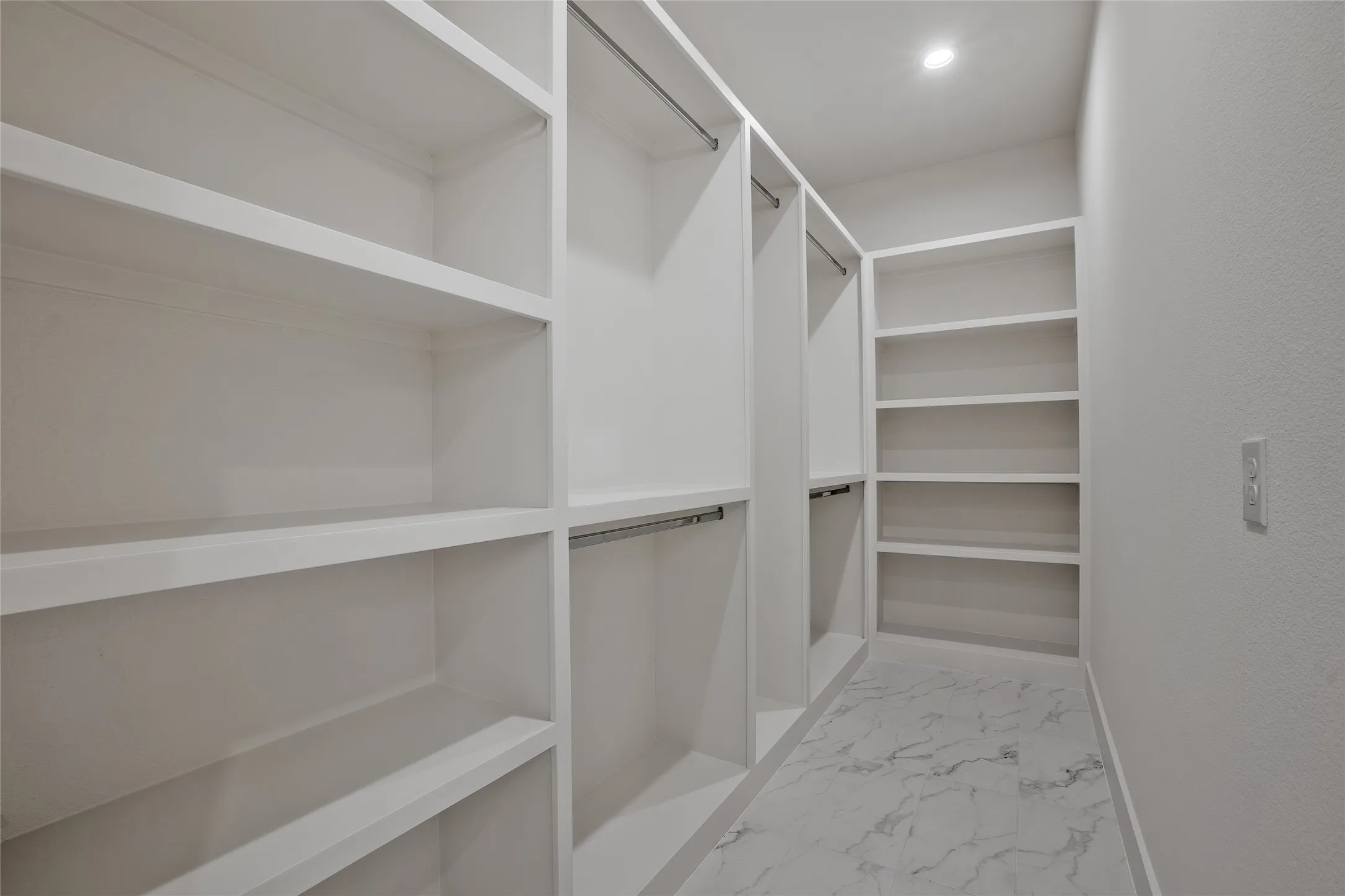 Walk-in closet featuring built-in white shelving, hanging rods, recessed lighting, and white tile flooring with grey veining