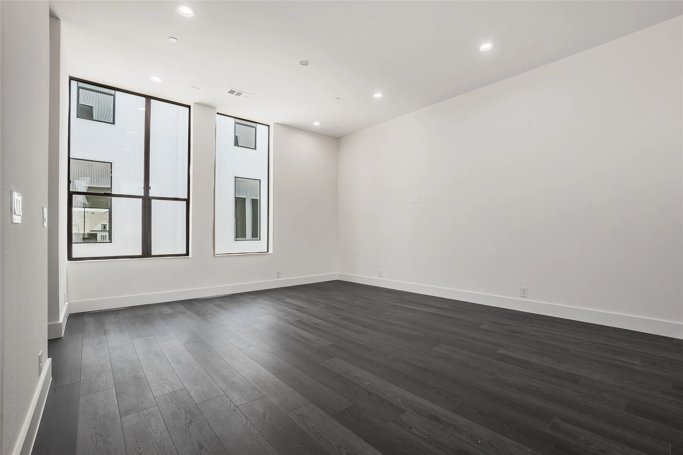 Spacious room featuring dark wood-finish flooring, contemporary large black-framed windows, recessed lighting, white baseboards, and a clean white wall finish