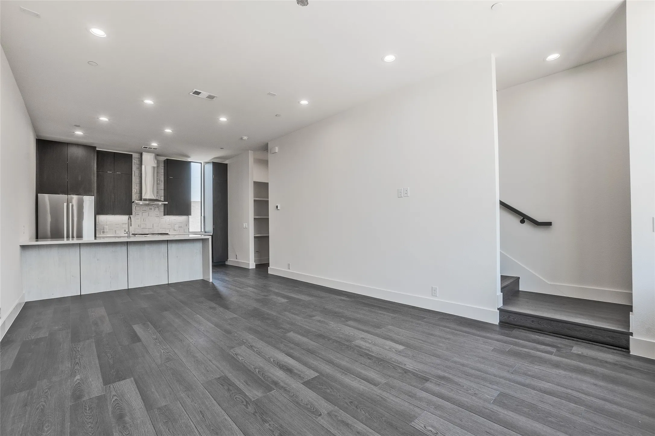 Open-concept living area with wood-finish flooring, recessed lighting, and a contemporary kitchen featuring stainless steel appliances, a stone backsplash, and dark-toned cabinetry