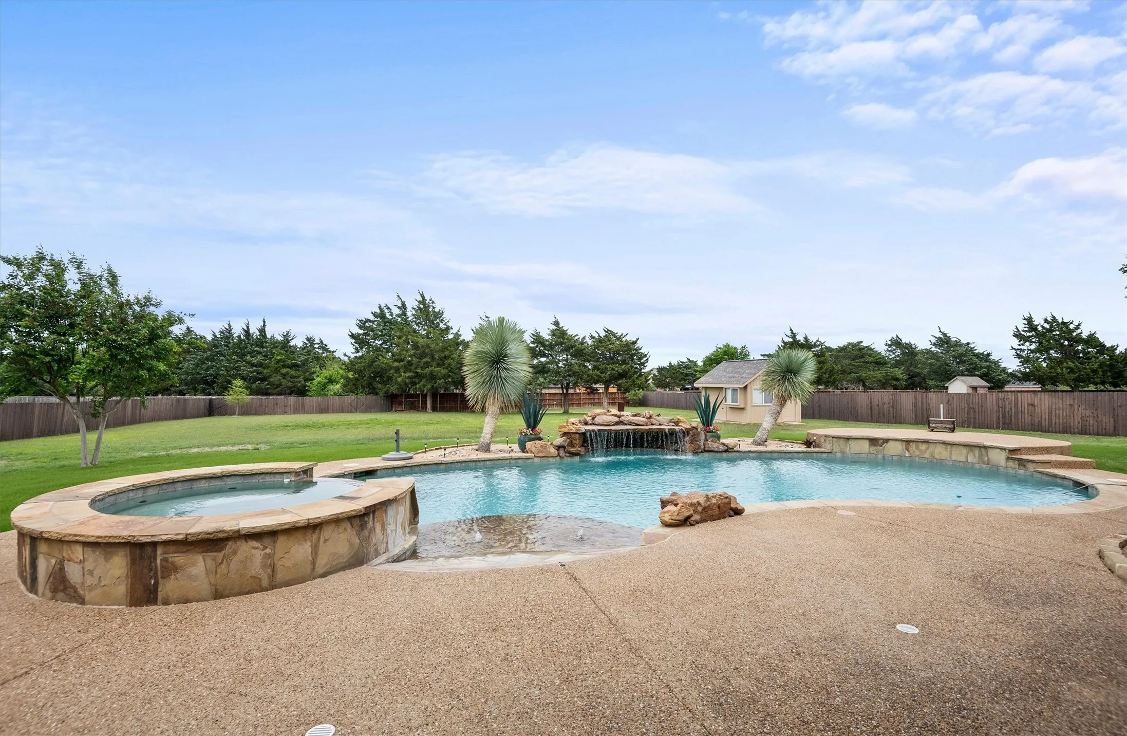 Expansive backyard featuring a large swimming pool with a rock waterfall, an integrated spa with stone surround, a textured concrete patio, a detached storage shed, and a privacy fence
