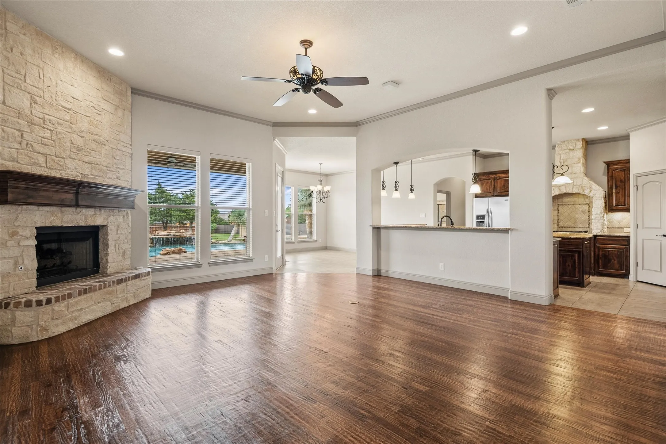 Spacious living area featuring wood-finish flooring, a stone fireplace with a substantial wood mantel, and multiple windows offering views of the exterior pool
