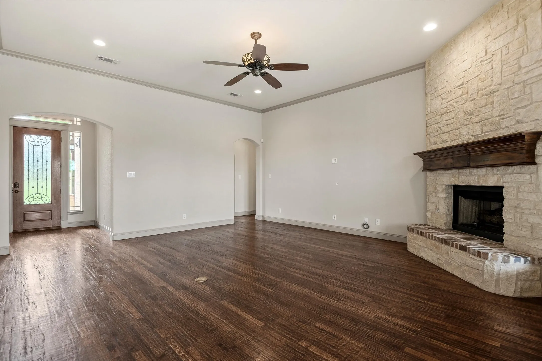 Spacious living area featuring rich wood-finish flooring, a stone-clad fireplace with a decorative wood mantle, recessed lighting, and a ceiling fan