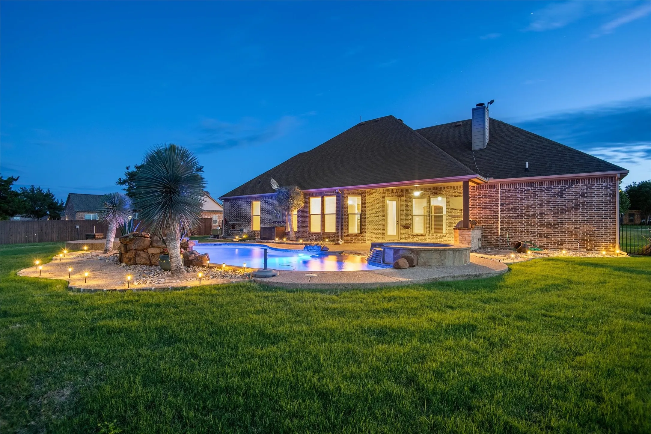 Expansive backyard featuring a custom pool with integrated spa, concrete patio surround, lush green lawn, and illuminated landscaping