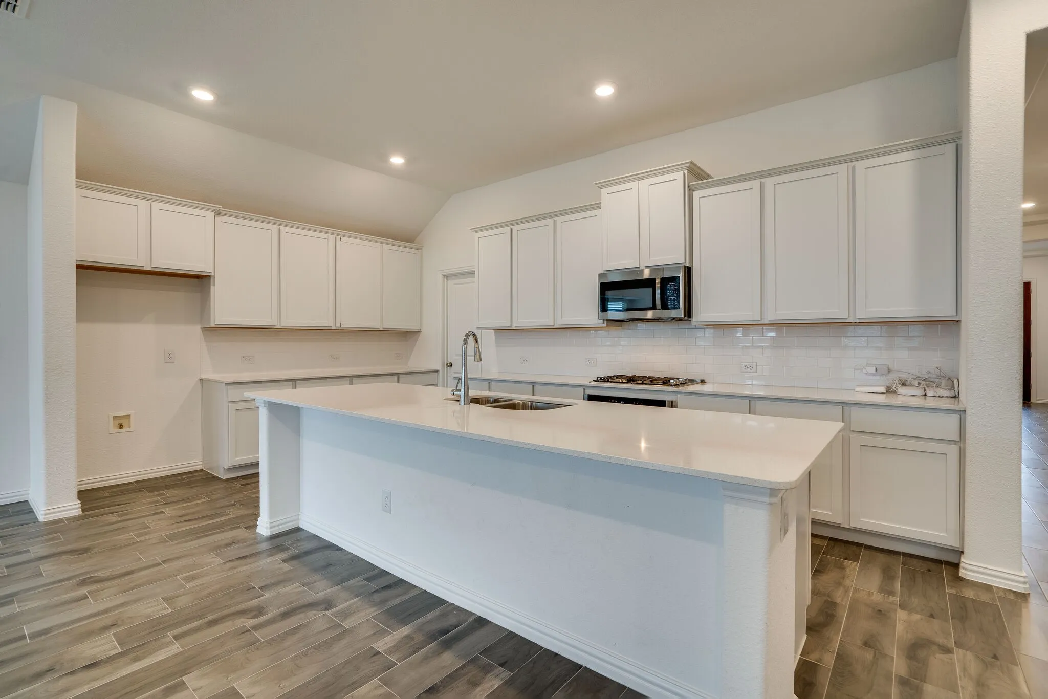 Spacious kitchen featuring a large central island with an undermount sink, extensive white cabinetry, light-toned countertops, subway tile backsplash, and wood-finish flooring