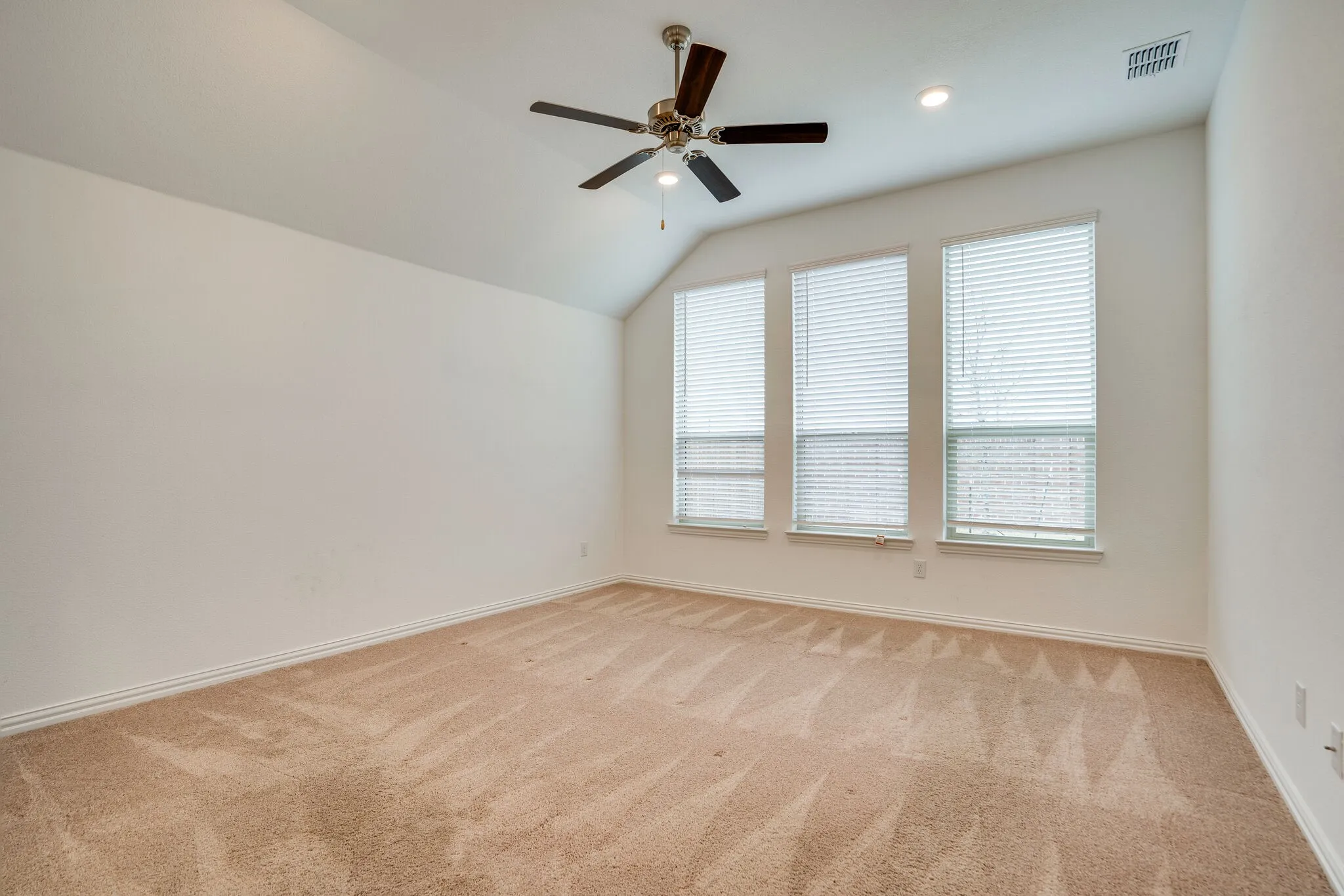 Spacious room featuring vaulted ceilings, plush carpet flooring, three large windows with blinds, a ceiling fan, and recessed lighting