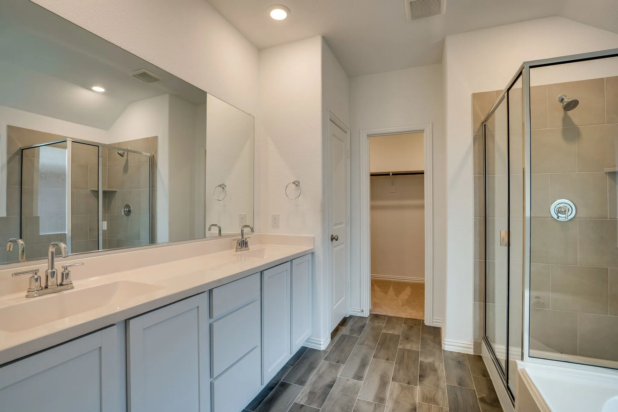 Bathroom featuring a dual vanity with light cabinetry and white countertops, a frameless glass shower enclosure with tiled walls, wood-look flooring, and a recessed ceiling light