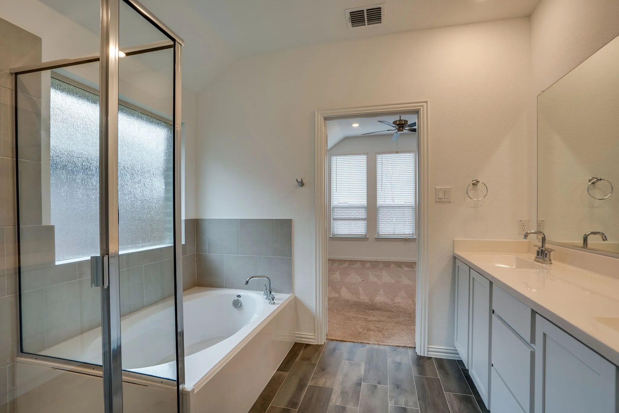 Wood-finish flooring bathroom featuring a built-in tub, tiled shower with glass enclosure, dual vanity with undermount sinks, and a ceiling fan visible through the doorway