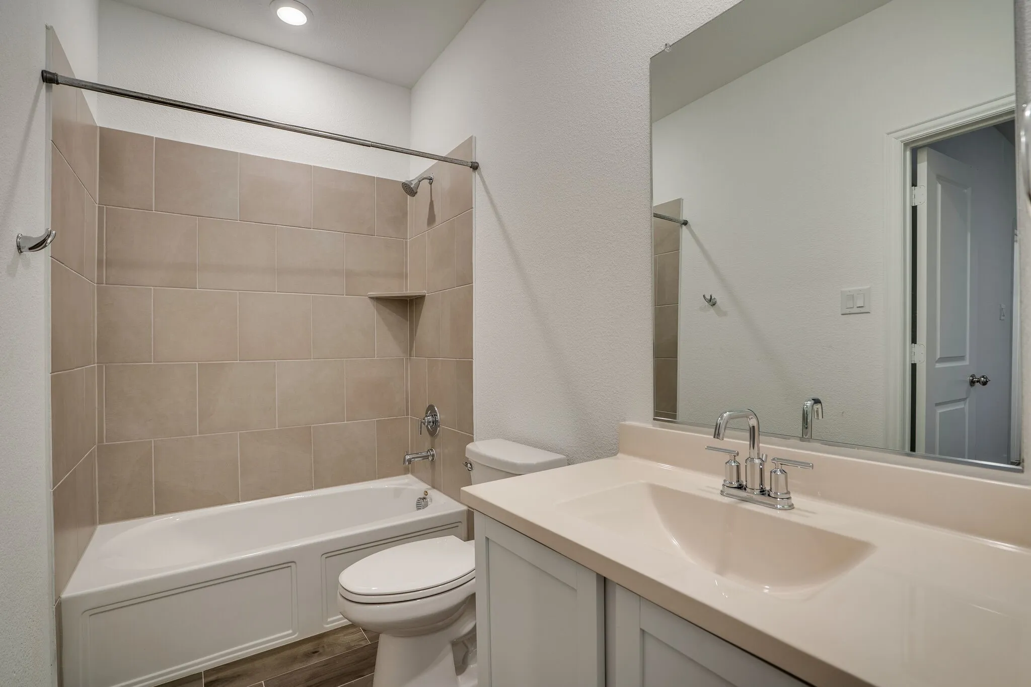 Full bathroom featuring a white porcelain tub and shower combination with tan tile surround, a built-in shower shelf, a vanity with a light-colored countertop and integrated sink, and wood-finish flooring