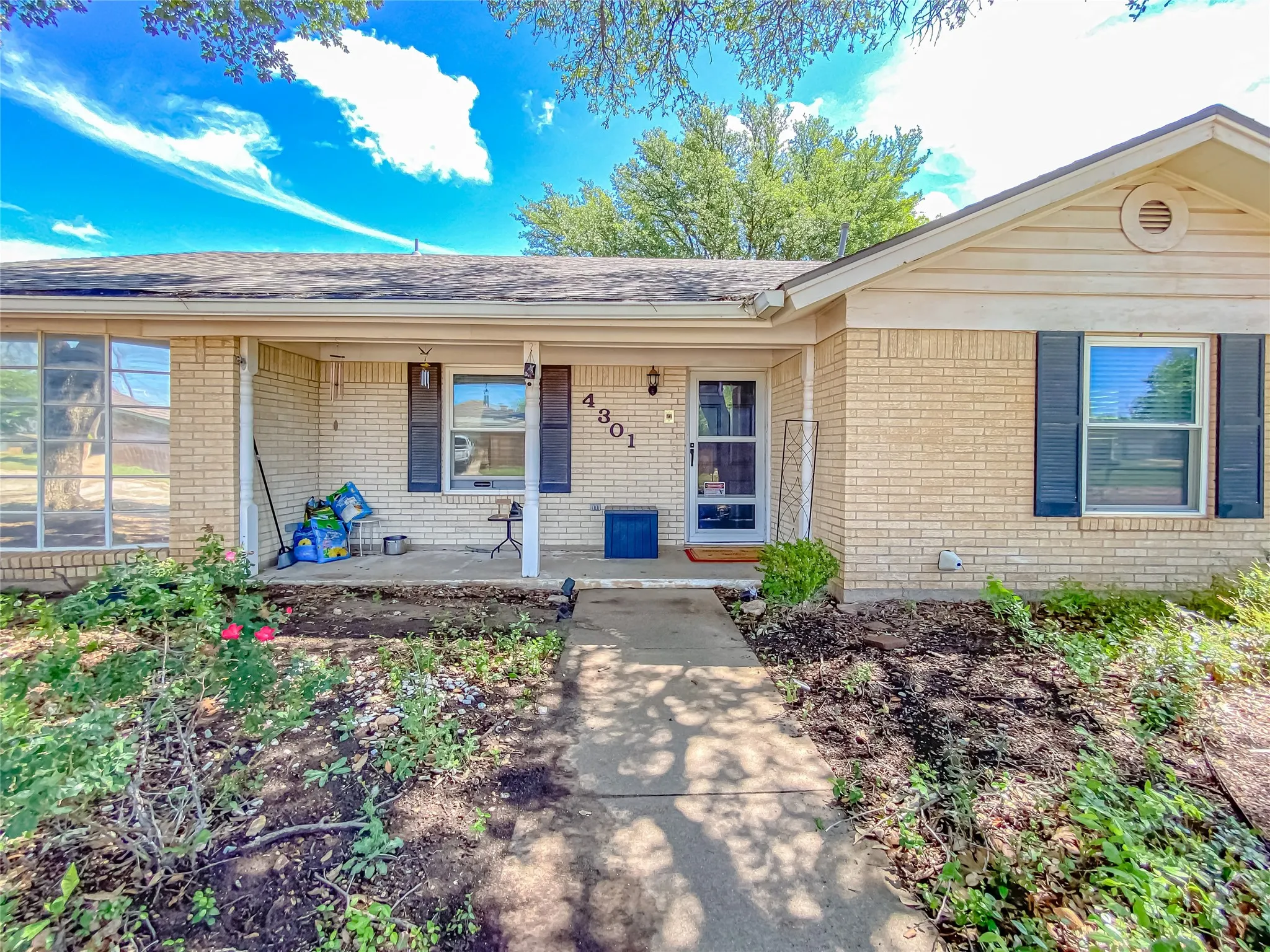 Light brick exterior with contrasting dark window shutters, an inviting covered front porch, and an established front yard featuring garden beds and mature trees