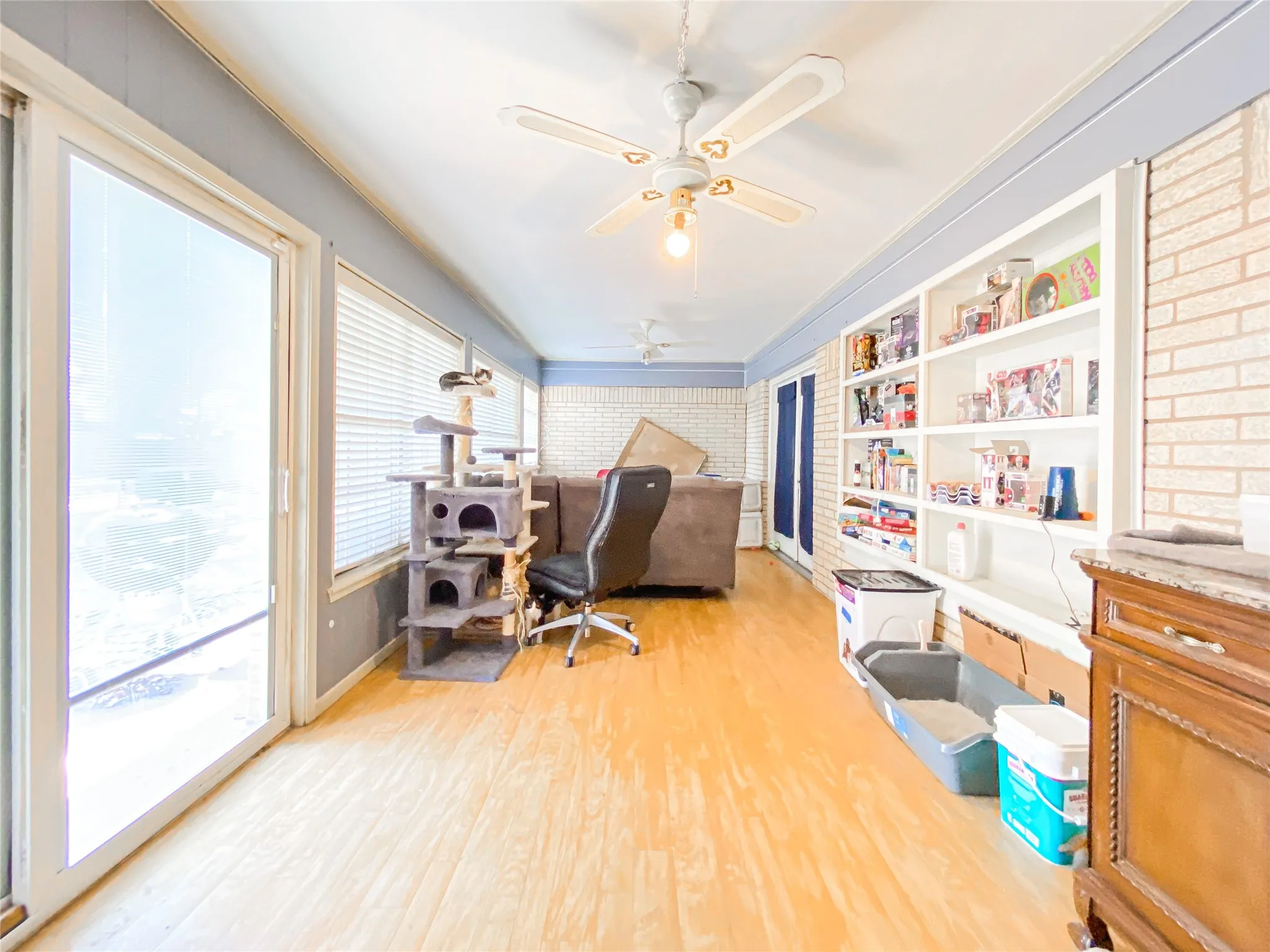 Bright room featuring wood-finish flooring, a full wall of built-in shelving, white brick accent walls, and ceiling fans