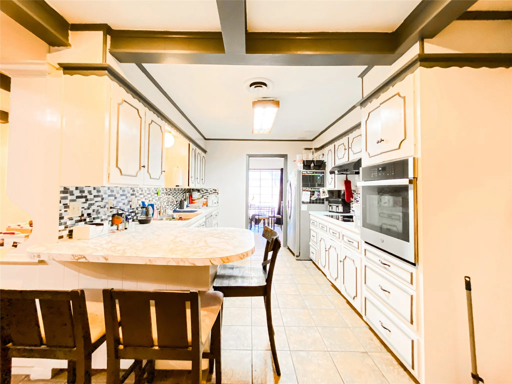 Galley kitchen featuring white cabinetry with gold trim, a built-in oven, tile flooring, a multi-tone tile backsplash, and a breakfast bar with a stone-finish countertop