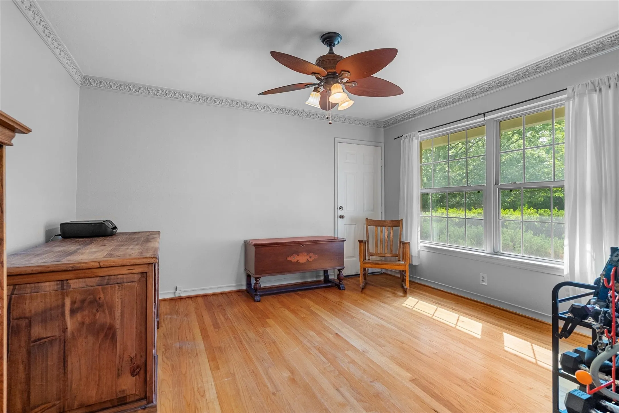 Hardwood flooring throughout this versatile room, featuring a white paneled door, a multi-pane window, decorative crown molding, and a ceiling fan with integrated lighting