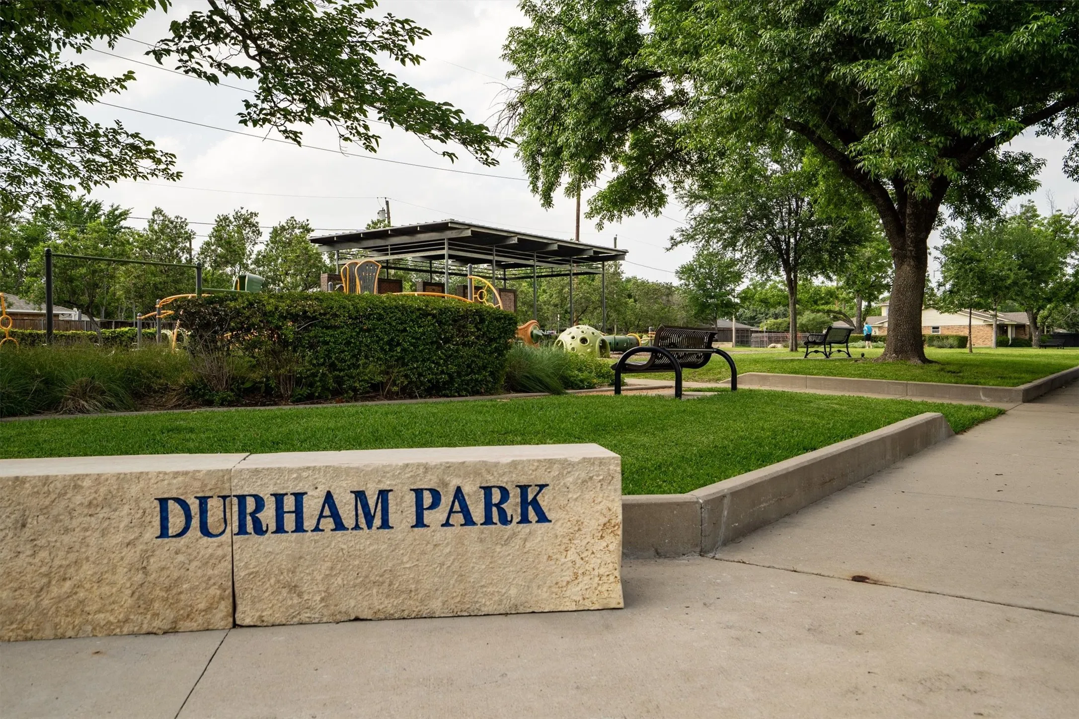 Durham Park stone signage marking entry to green space featuring mature trees and a shaded playground area