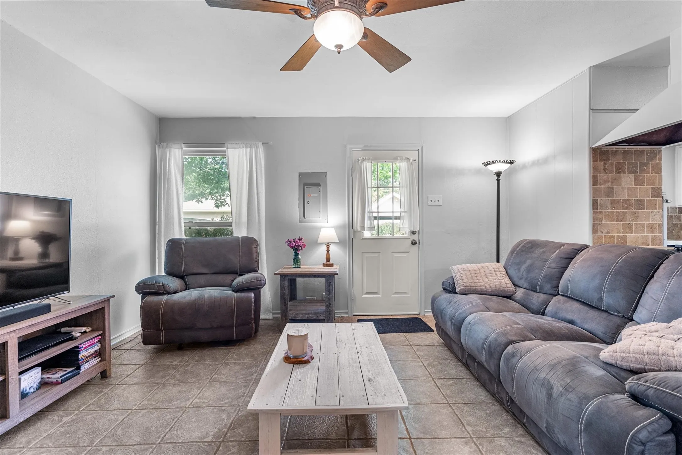 Spacious living area featuring tile flooring, a ceiling fan with integrated lighting, a window with white curtains, a paneled door with a glass insert, and light gray wall finishes