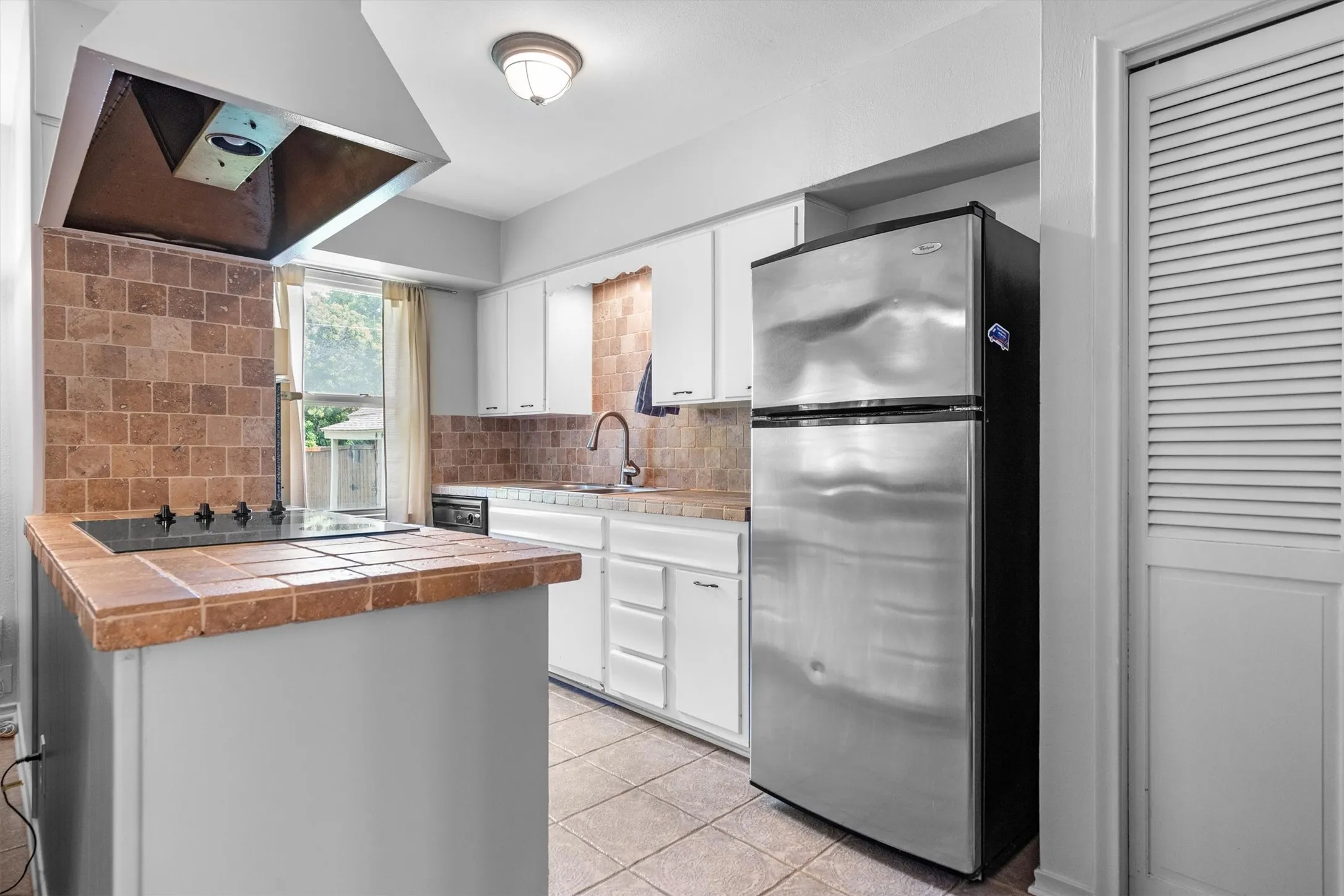 Kitchen featuring tile flooring, white cabinetry, a built-in cooktop, tile countertops, and a stainless steel​​‌​​​​‌​​‌‌​‌‌​​​‌‌​‌​‌​‌​​​‌​​ refrigerator