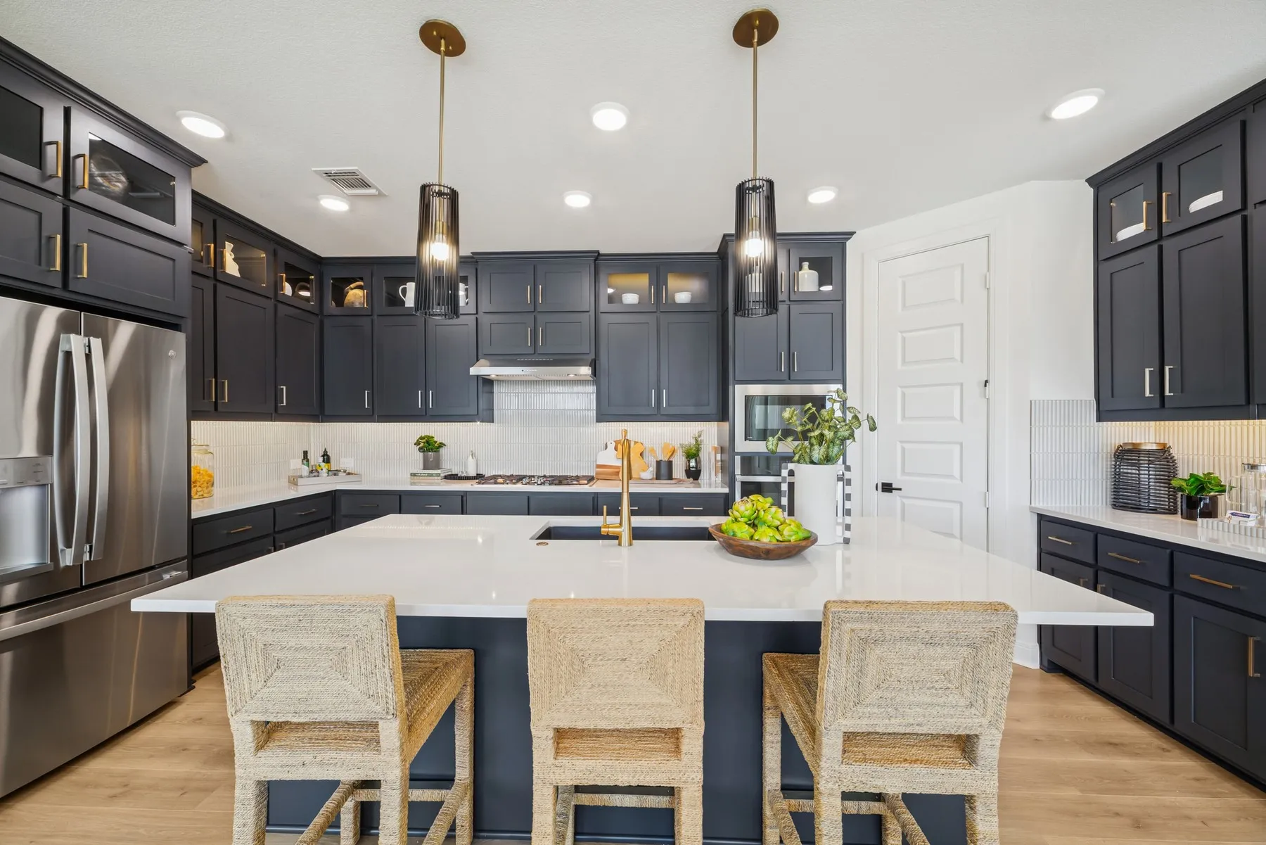 Modern kitchen with dark cabinetry, stainless steel appliances, a large central island with seating, pendant lighting, and light wood-finish flooring