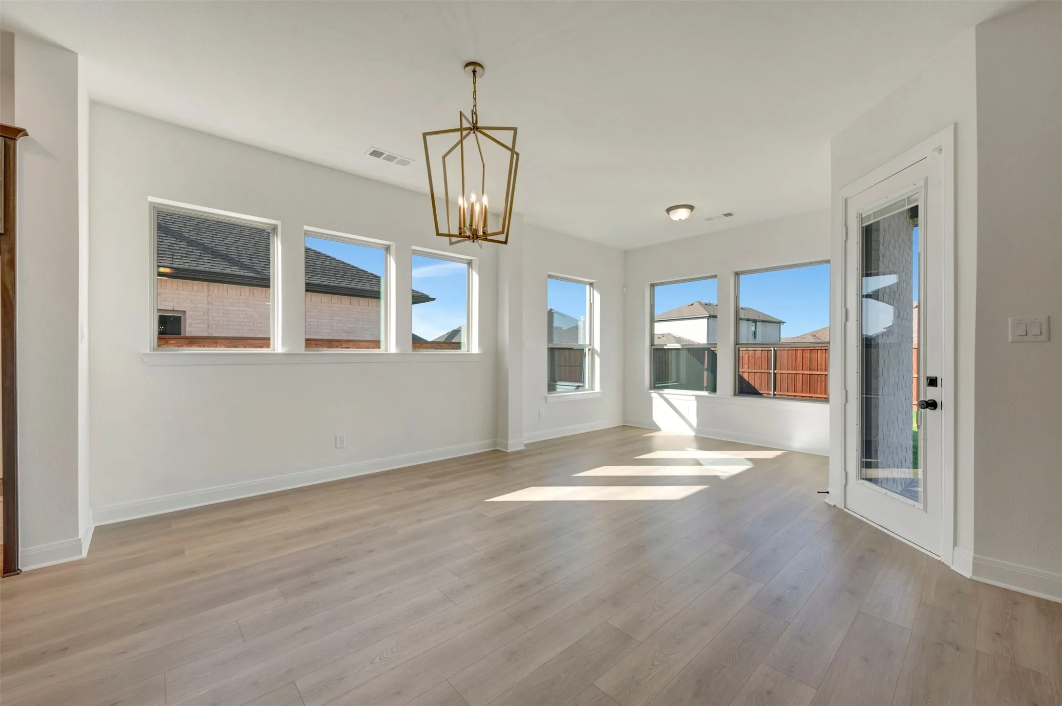 Spacious room with wood-finish flooring, multiple windows, a geometric chandelier, a ceiling-mounted light fixture, and a glass-paneled door