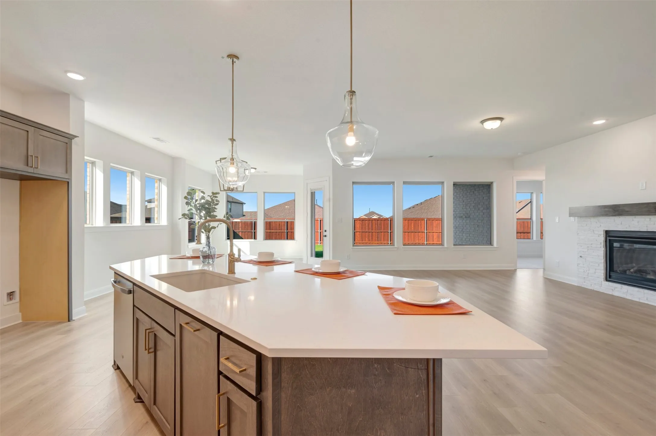 Open-concept interior featuring light wood-finish flooring, a kitchen island with integrated sink, pendant lighting, and a fireplace with a tile surround and wood mantel