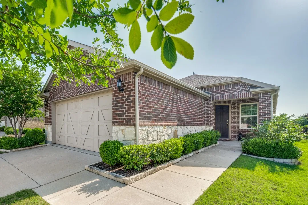 Exterior featuring a brick facade with stone accents, an attached garage with a panel door, a paved driveway, a concrete walkway, and landscaped shrubbery