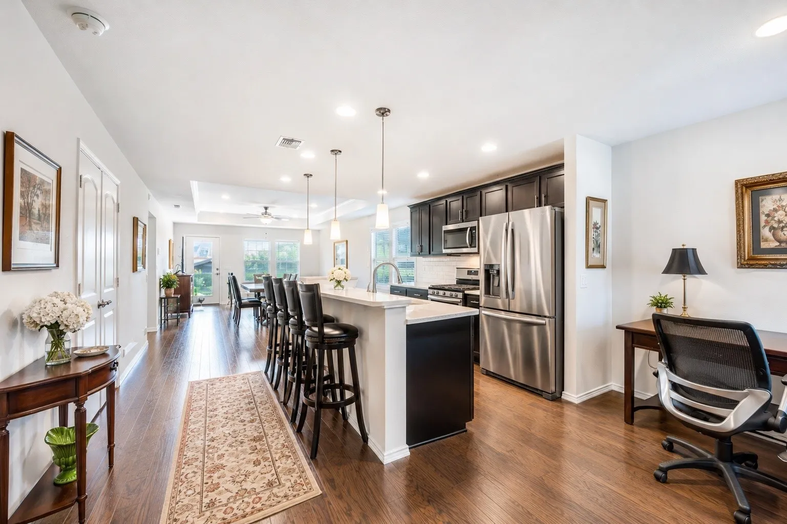 Open-concept living area featuring rich wood-finish flooring, a kitchen island with pendant lighting, dark wood cabinetry, stainless steel appliances, and a dedicated desk area