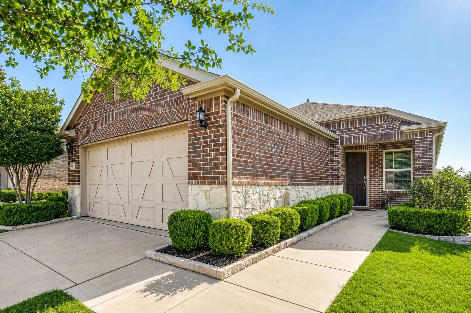 Brick and stone exterior featuring an attached garage, landscaped front yard with mature shrubbery, a concrete driveway, and a covered entry