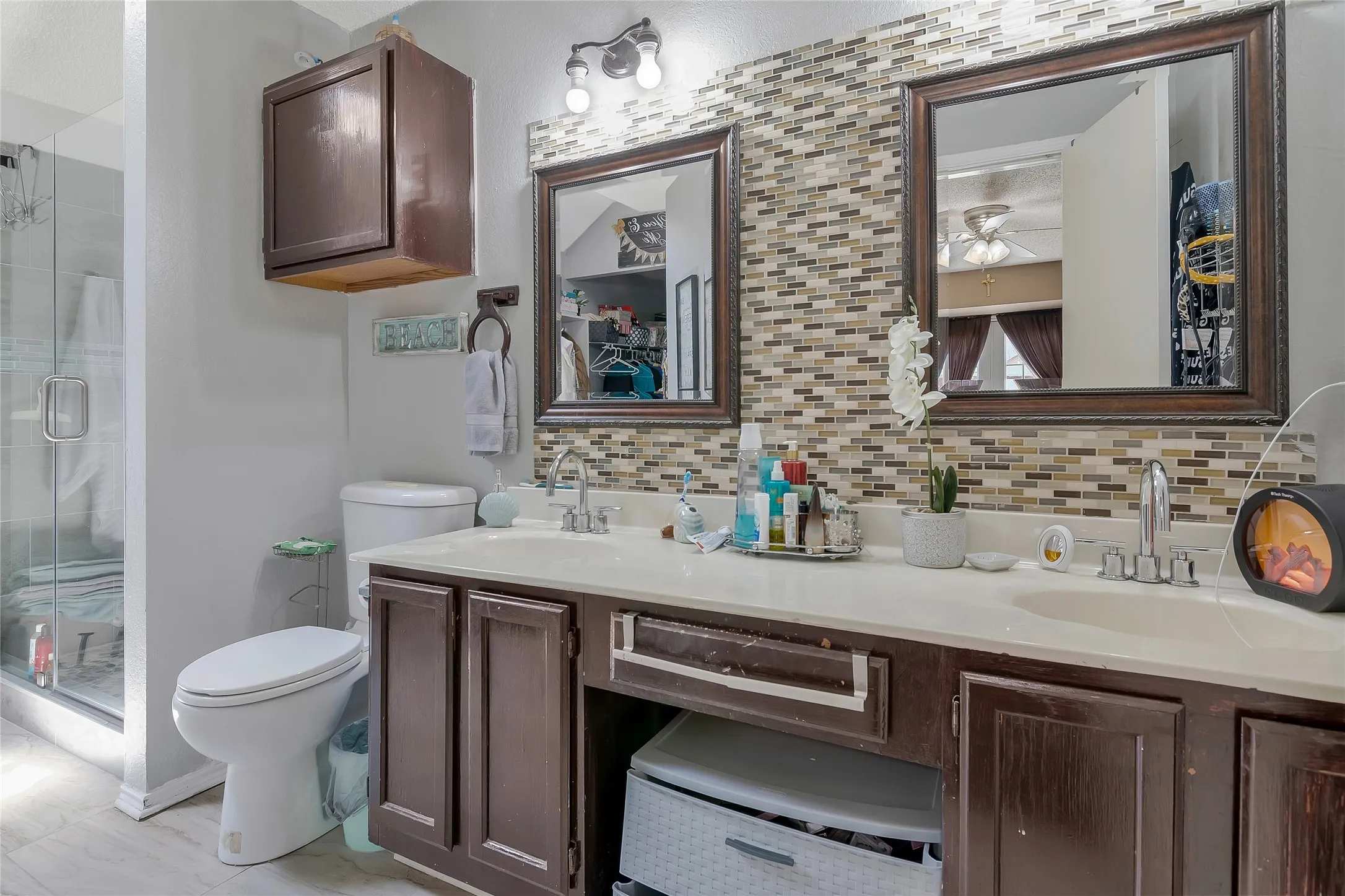 Bathroom with backsplash, a shower with shower door, toilet, ceiling fan, and dual vanity