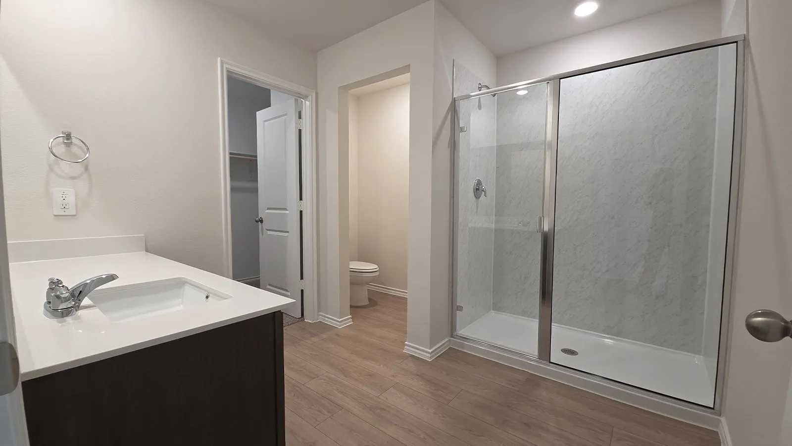 Spacious bathroom with wood-finish flooring, a contemporary vanity featuring a white countertop and integrated sink, and a glass-enclosed shower with subway tile surround