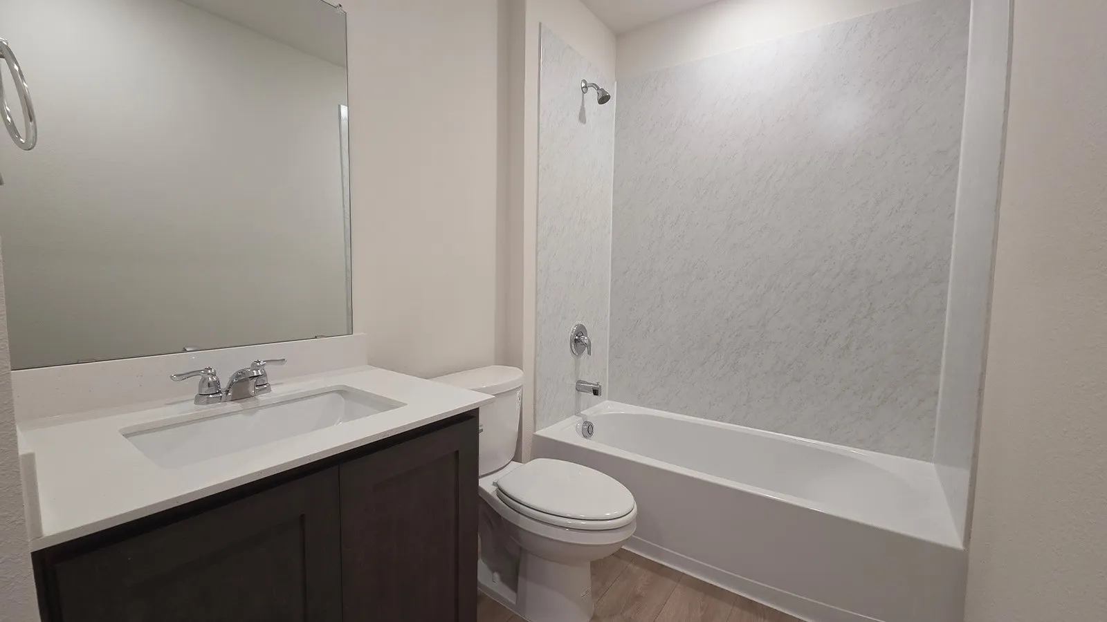 Bathroom featuring a dark wood-finish vanity with a white countertop and integrated sink, a large wall-mounted mirror, a standard toilet, and a white bathtub with a textured surround