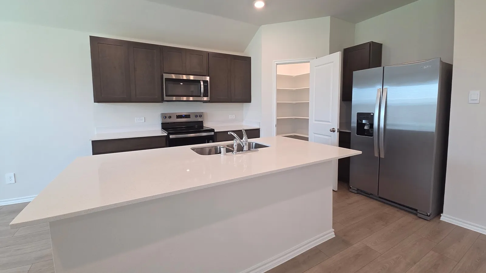 Modern kitchen featuring a large central island with an undermount sink, stainless steel appliances, dark wood-finish cabinetry, and light-toned countertops