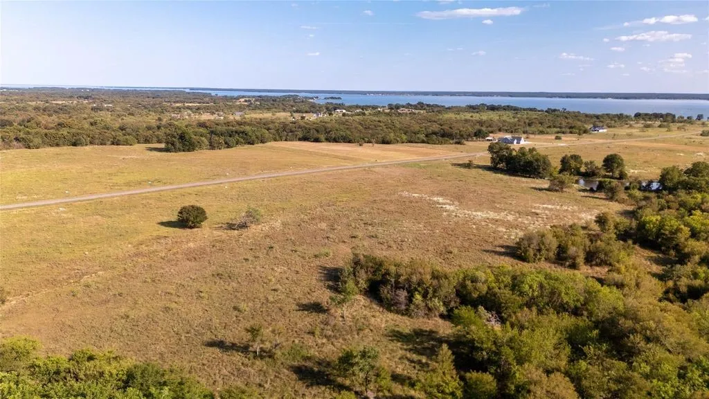 Expansive open land featuring a water body in the background, scattered trees, and a visible road