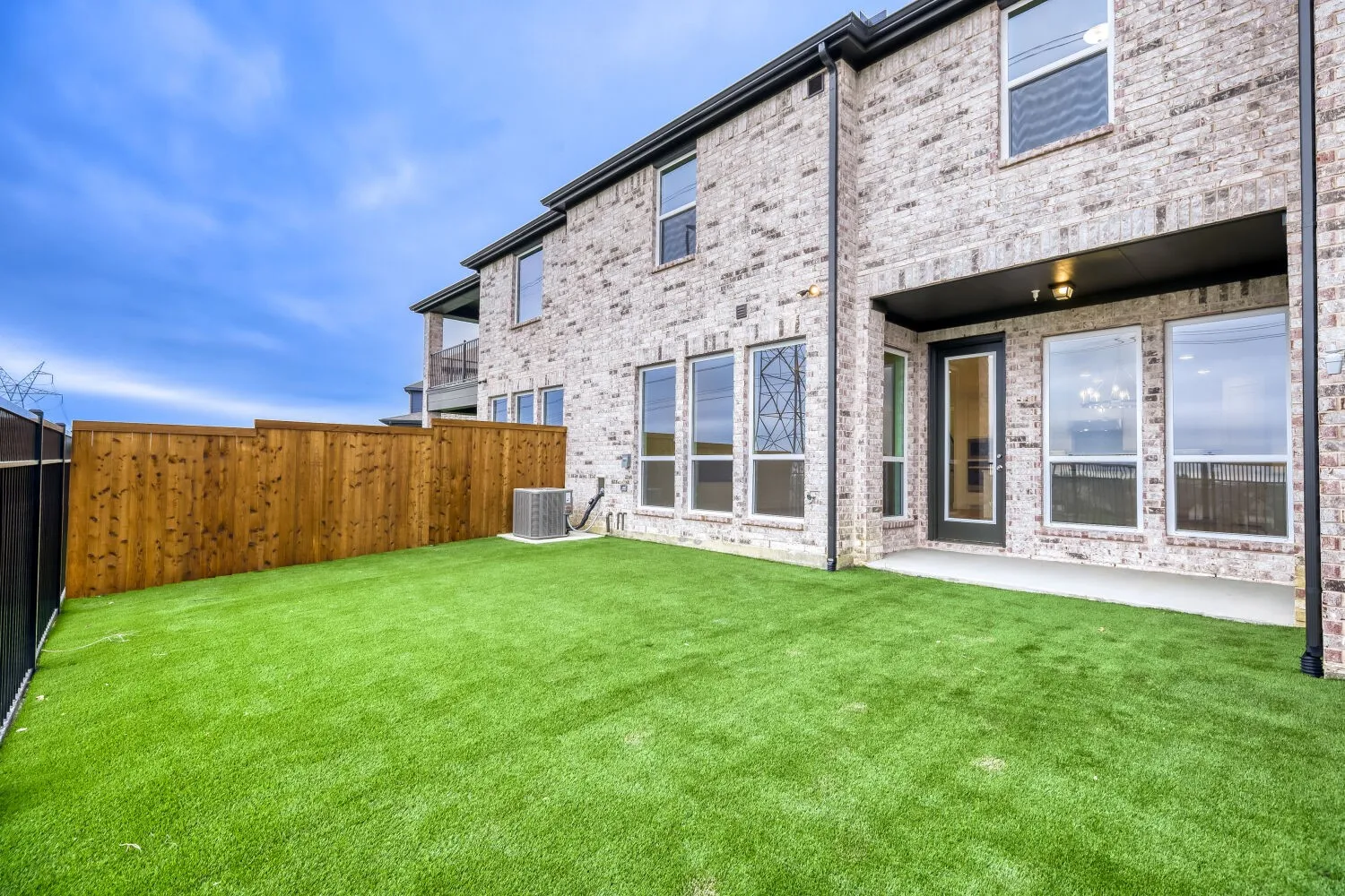 Expansive artificial turf backyard featuring a covered patio, extensive brick exterior, floor-to-ceiling windows, and a wood privacy fence