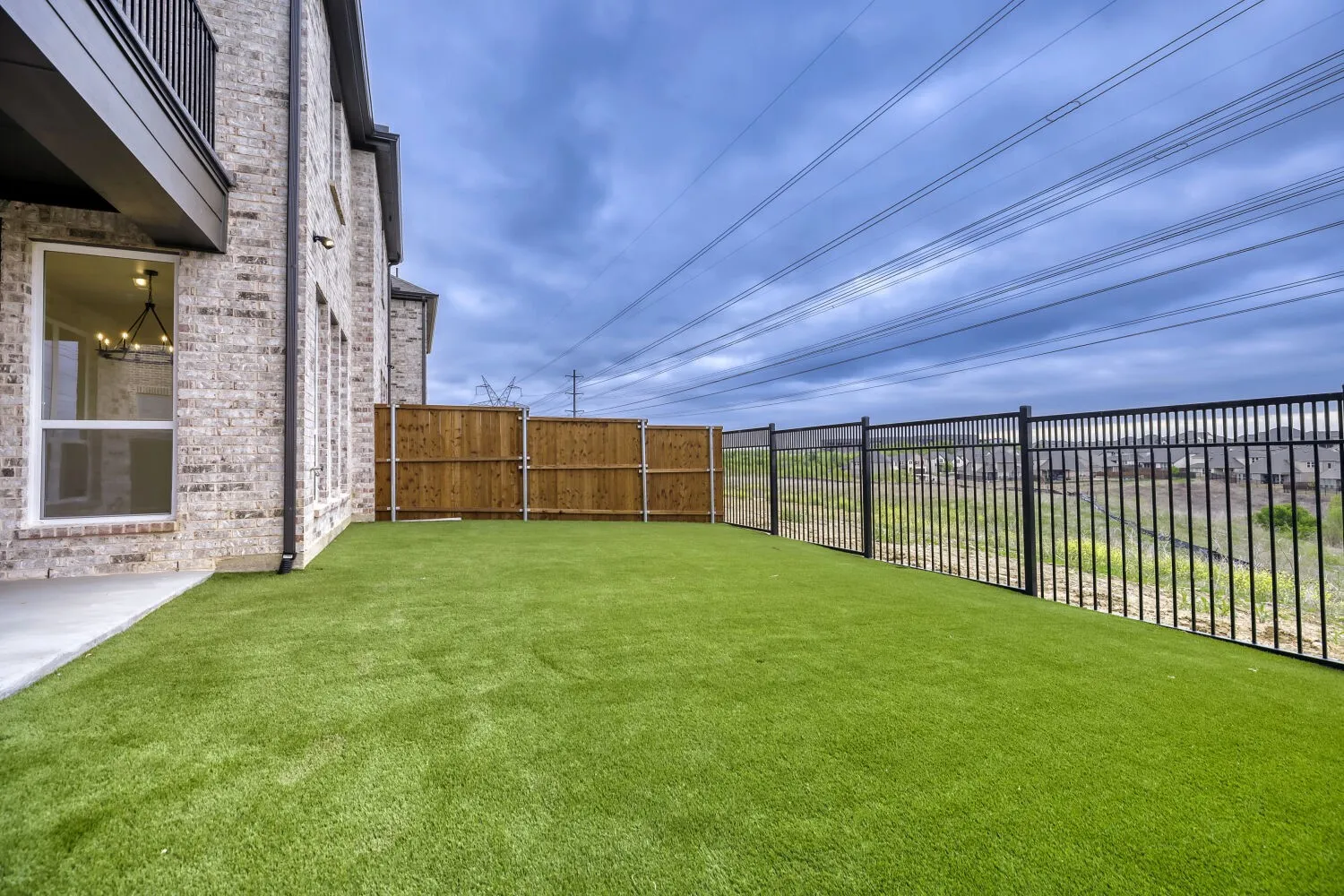 Expansive backyard featuring artificial turf, a concrete patio, a wooden privacy fence, and a black metal fence bordering the property