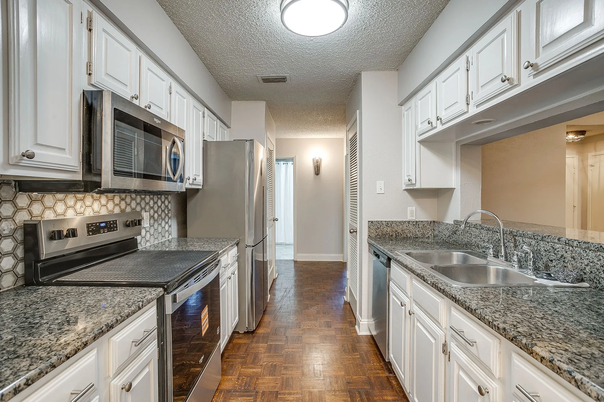 Galley kitchen featuring white cabinetry, stainless steel appliances, speckled stone countertops, and a honeycomb tile backsplash