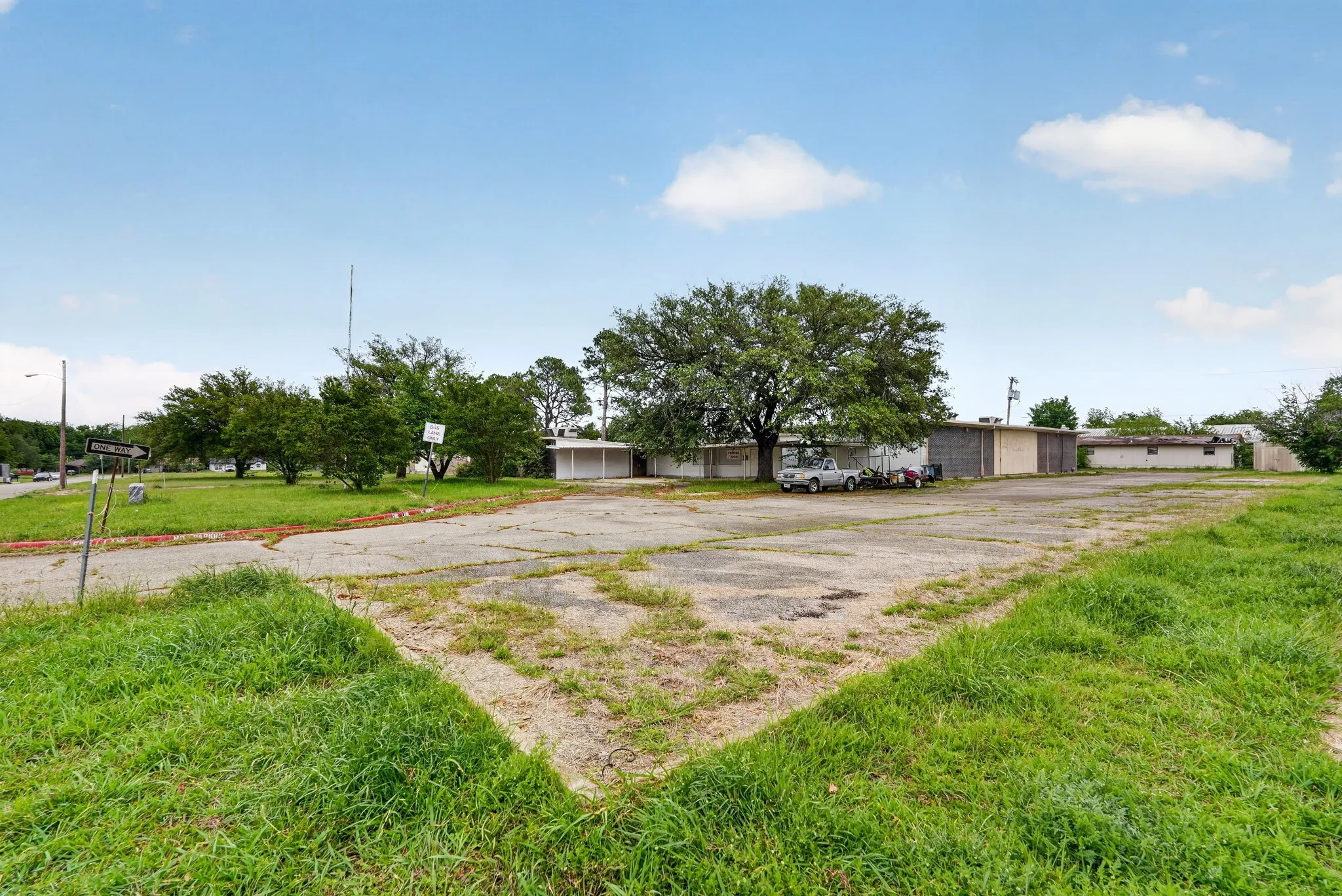 Expansive paved parking area with surrounding green space, mature trees, and single-story structures