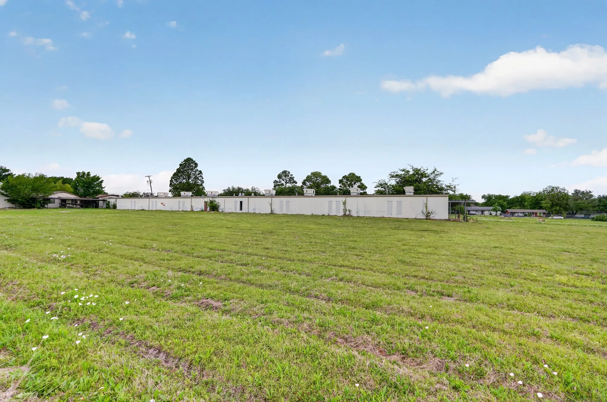 Expansive grassy lot with scattered wildflowers, featuring a long, single-story structure with multiple windows and a flat roofline