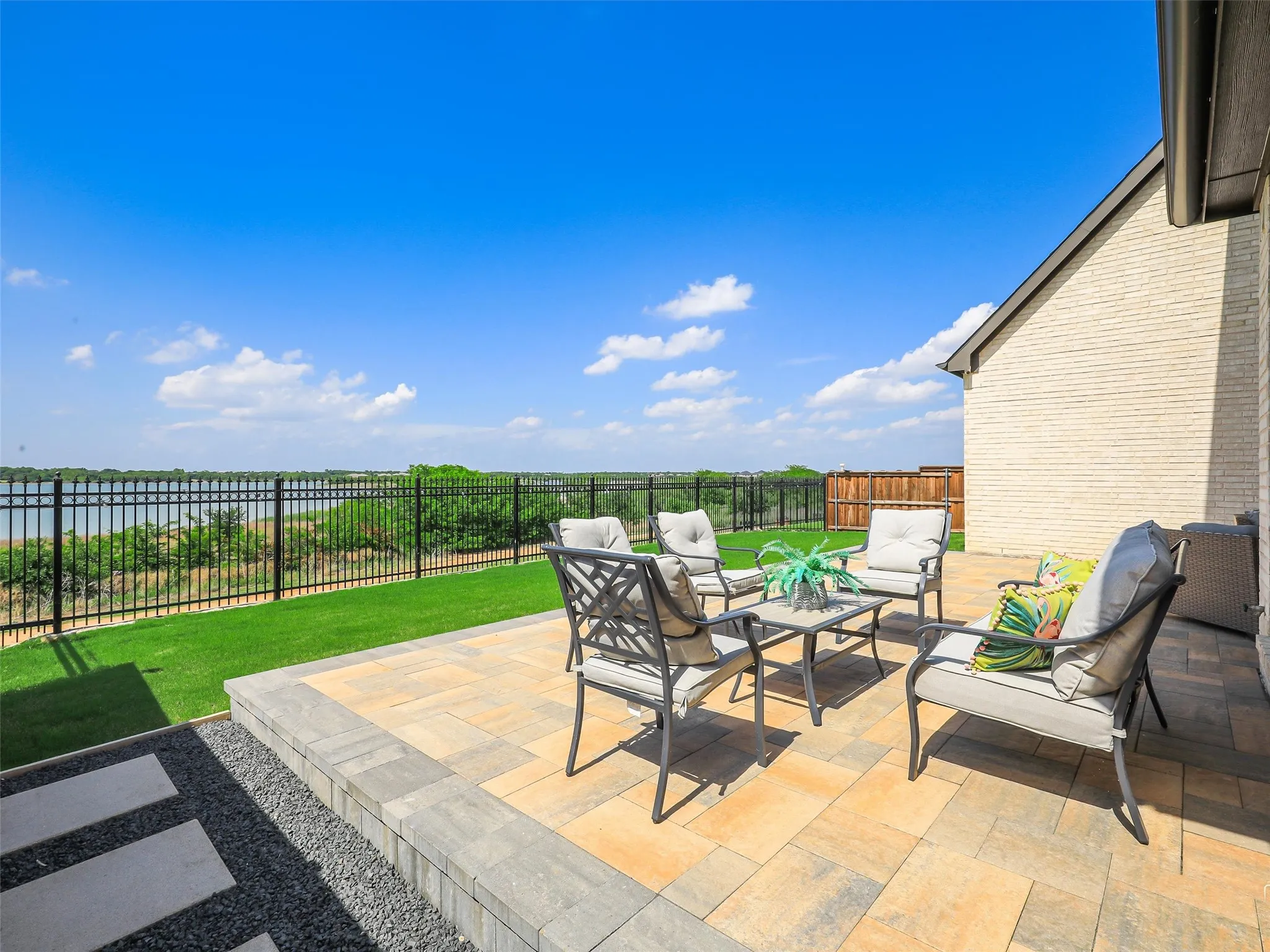 Expansive paved patio with a low retaining wall, overlooking a body of water and bordered by a black metal fence