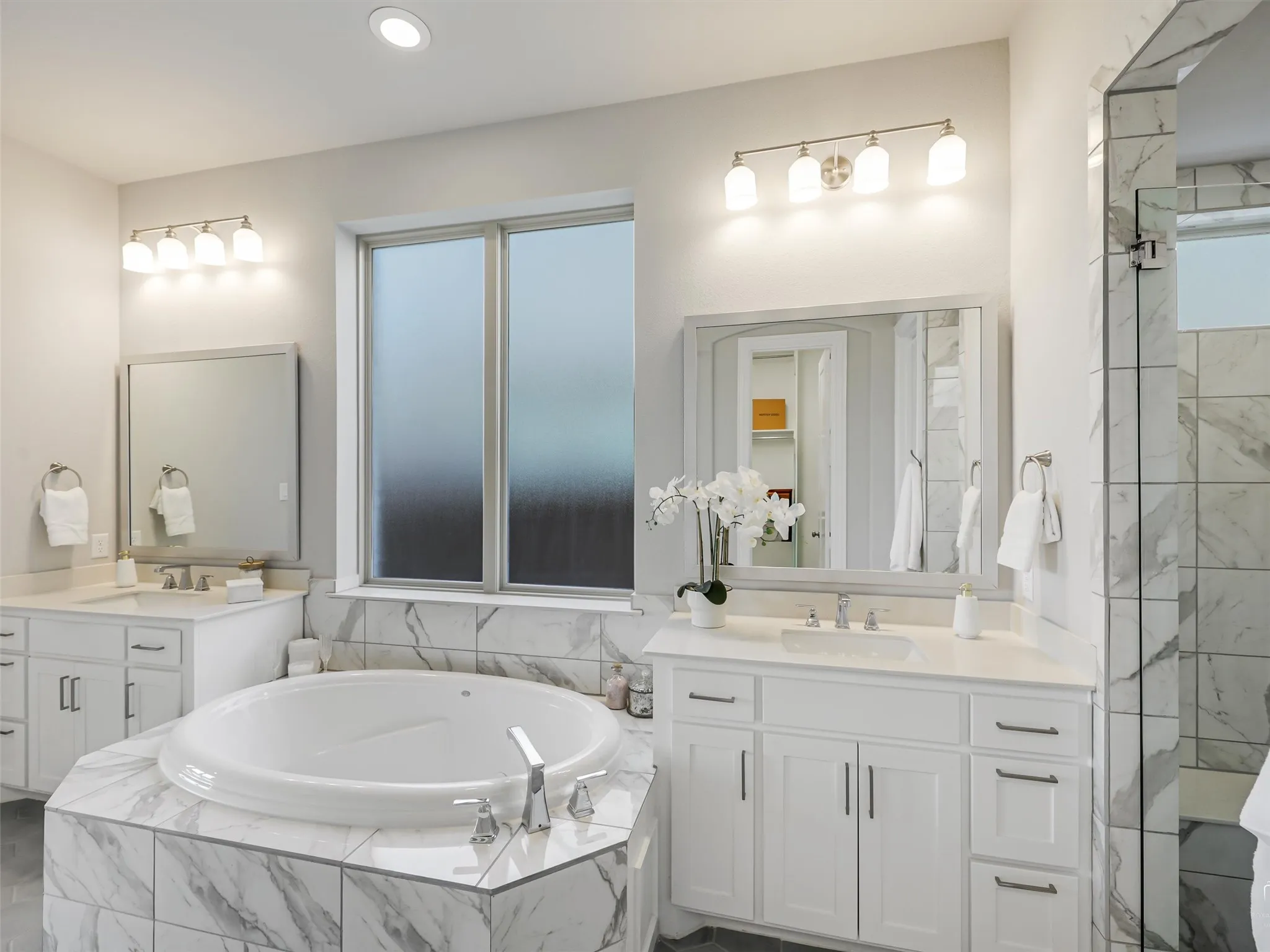 Built-in soaking tub surrounded by marble-look tile, dual vanities with white cabinetry, framed mirrors, and chrome fixtures, illuminated by overhead lighting and a frosted window