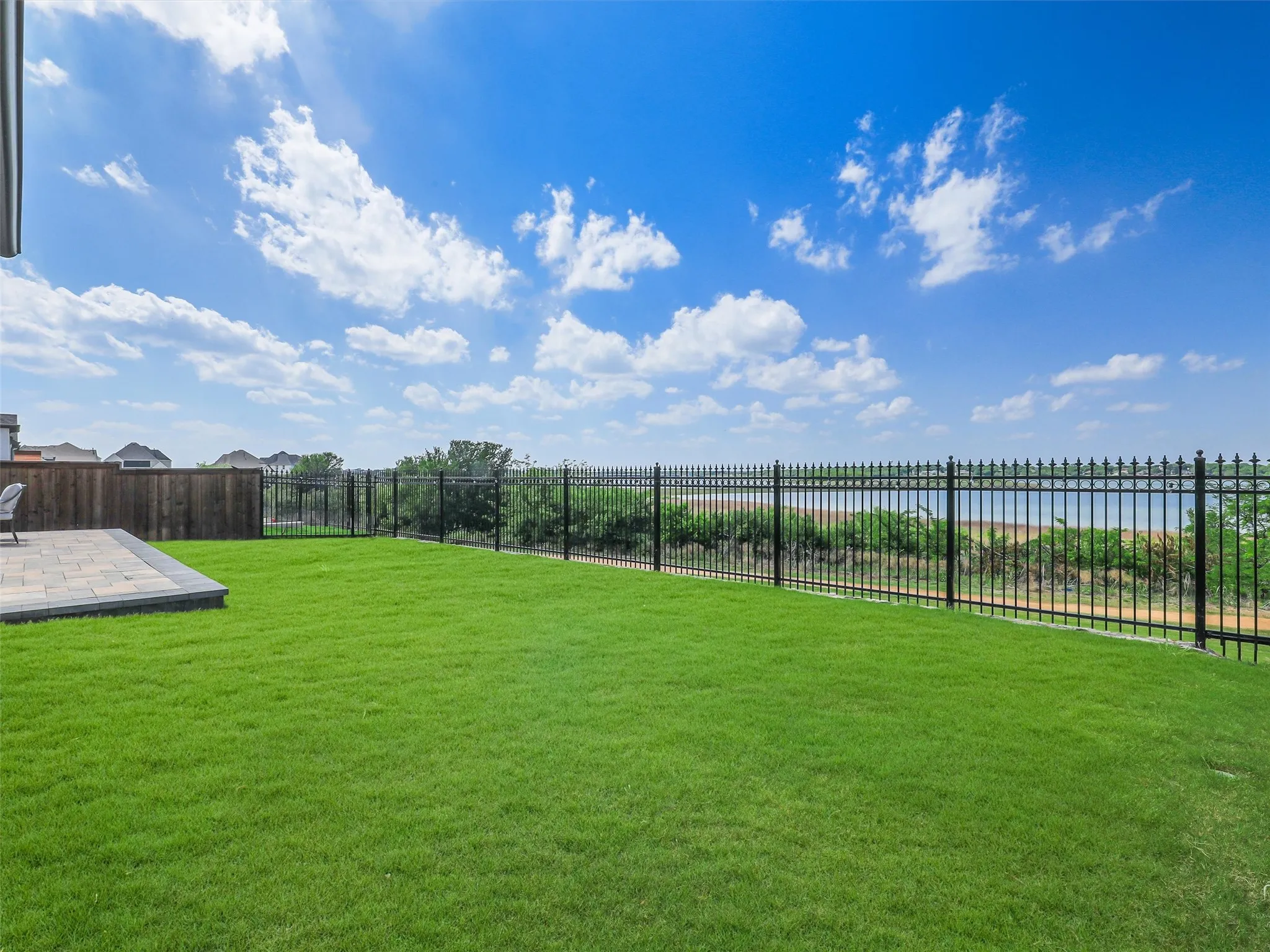 Expansive backyard featuring a green lawn, paver patio, black wrought iron fencing, and a lake backdrop
