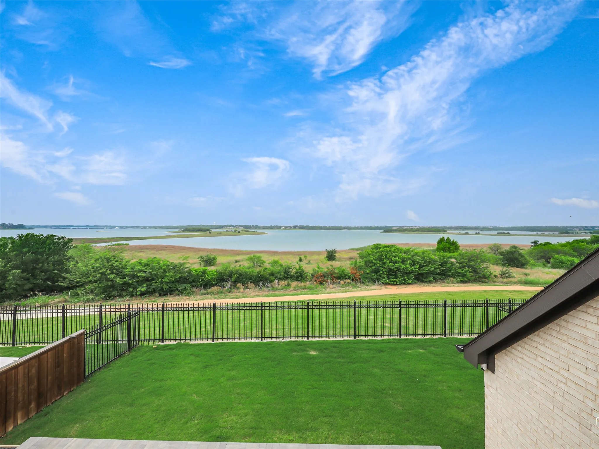Expansive green lawn bounded by a black metal fence, overlooking a substantial body of water with distant land features