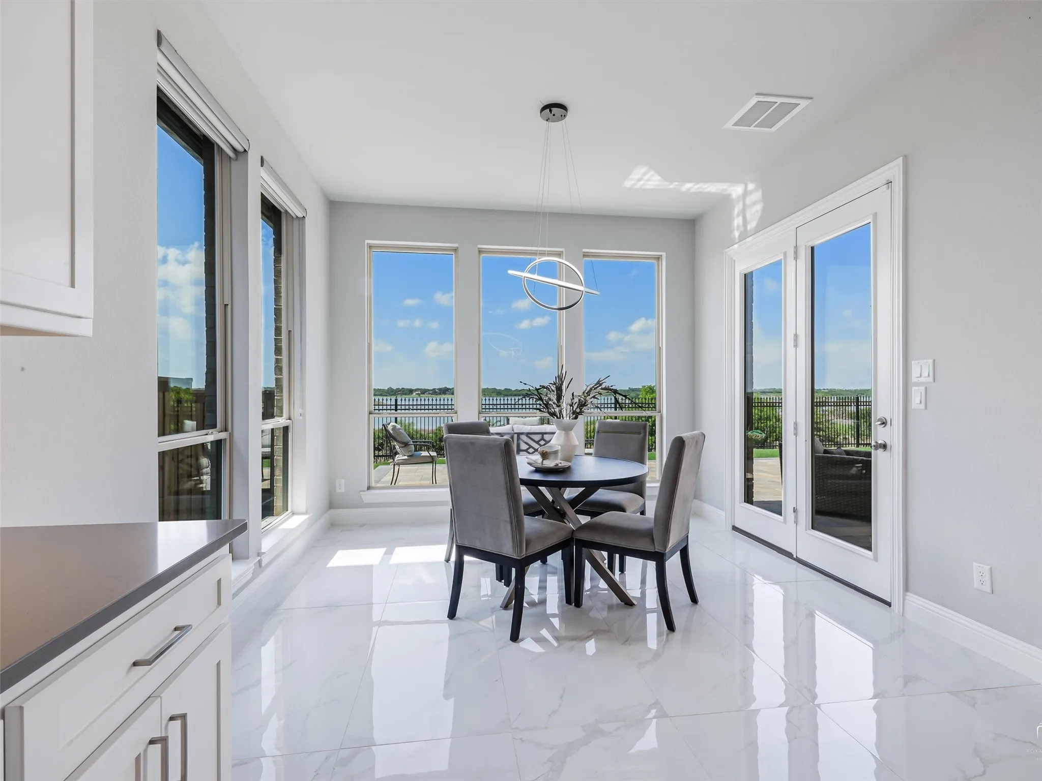 Bright dining area featuring large windows, a contemporary circular chandelier, white marbled tile flooring, and double glass-paneled doors opening to an outdoor space