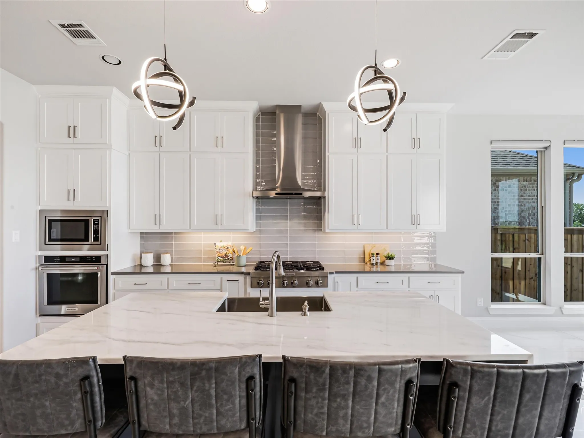 Kitchen featuring a large central island with a built-in sink, white cabinetry with contemporary hardware, stainless steel appliances, a tile backsplash, and two distinctive pendant light fixtures