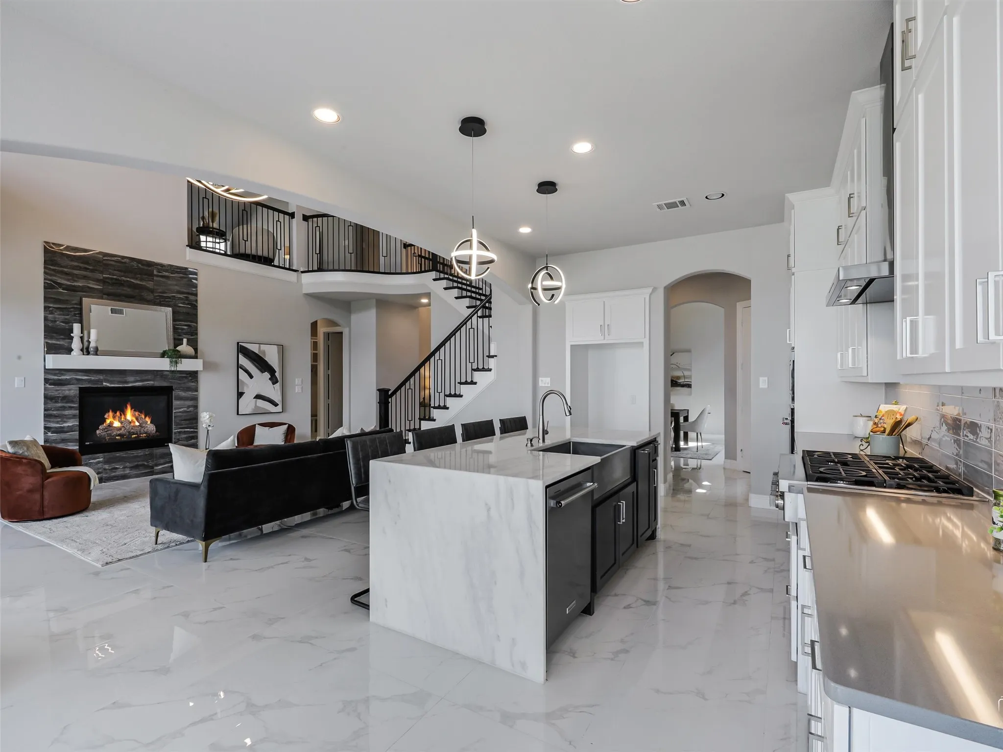 Open-concept living space featuring a modern kitchen island with waterfall countertop, a contemporary fireplace with a stone surround, a grand staircase with wrought iron balusters, white tile flooring, and integrated kitchen appliances