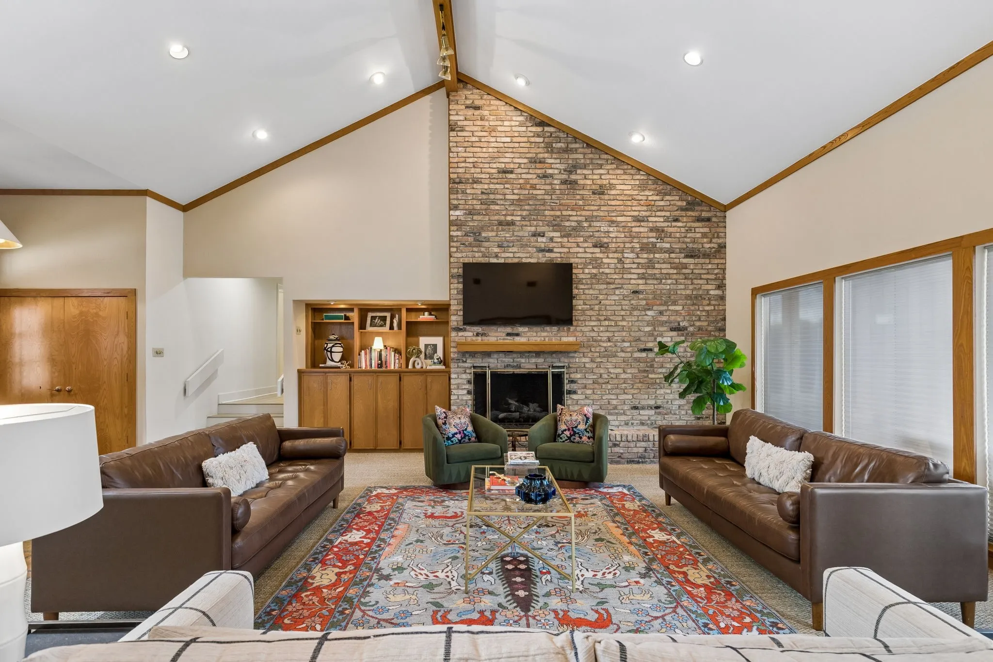 Vaulted ceilings with exposed wood beams, a prominent brick fireplace with a wood mantel, built-in shelving, recessed lighting, and large windows with horizontal blinds