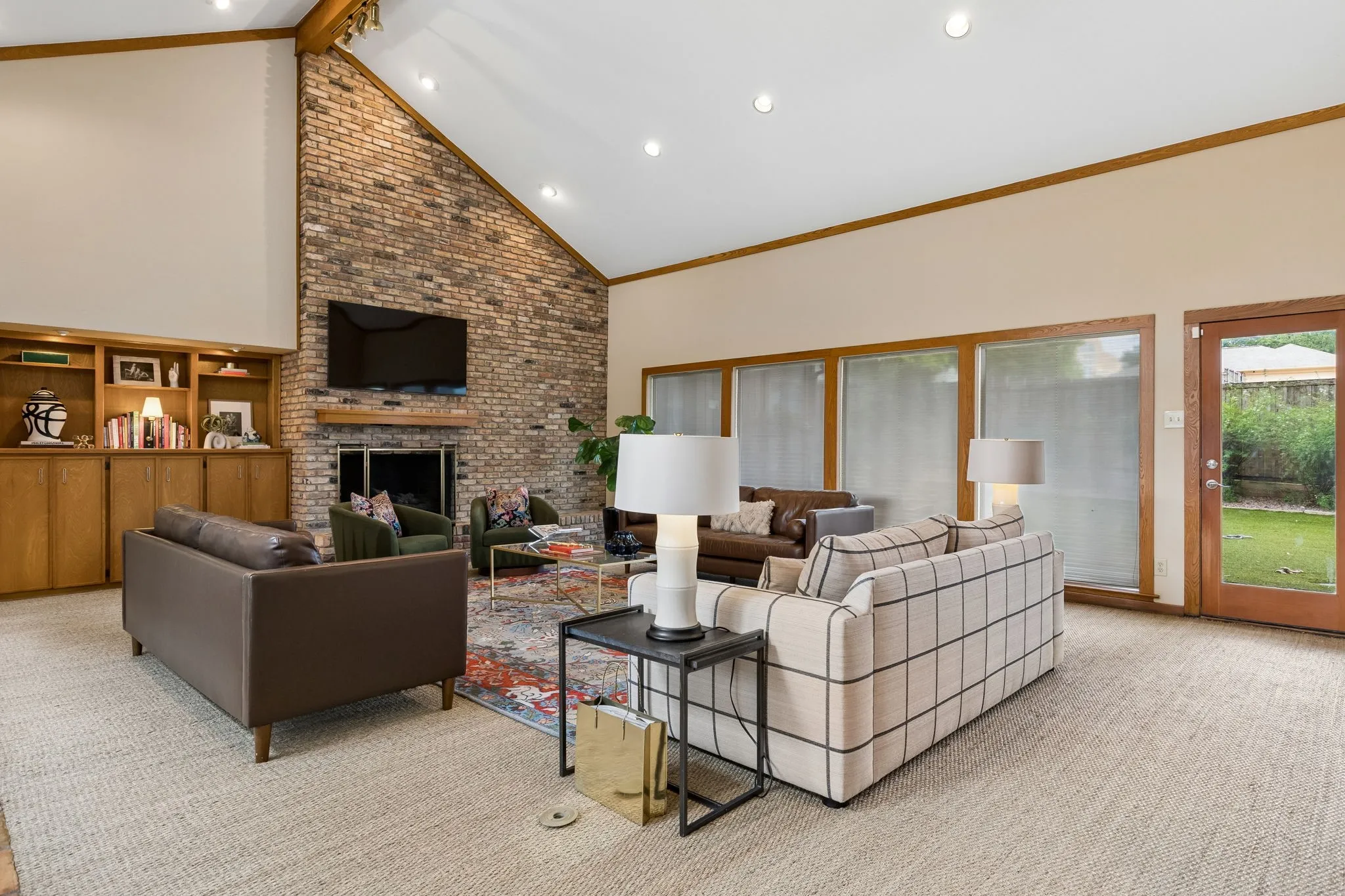 Vaulted ceiling living area featuring a brick fireplace, built-in cabinetry, recessed lighting, wood-finish window frames, and a glass-paneled door opening to the outdoors