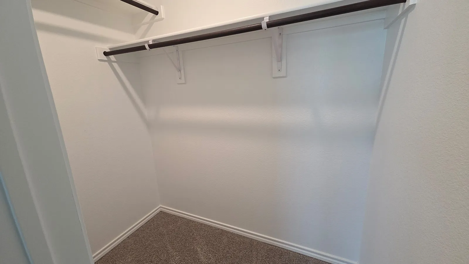 Walk-in closet featuring white painted walls, a built-in shelf, two dark-toned hanging rods, and gray carpet flooring